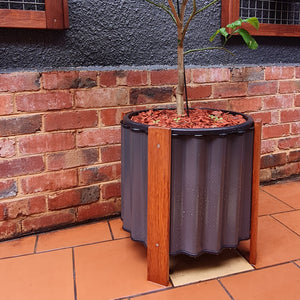A brown wooden planter box with a black plastic tub inside, placed on a tiled floor against a brick wall with a young plant inside the pot.